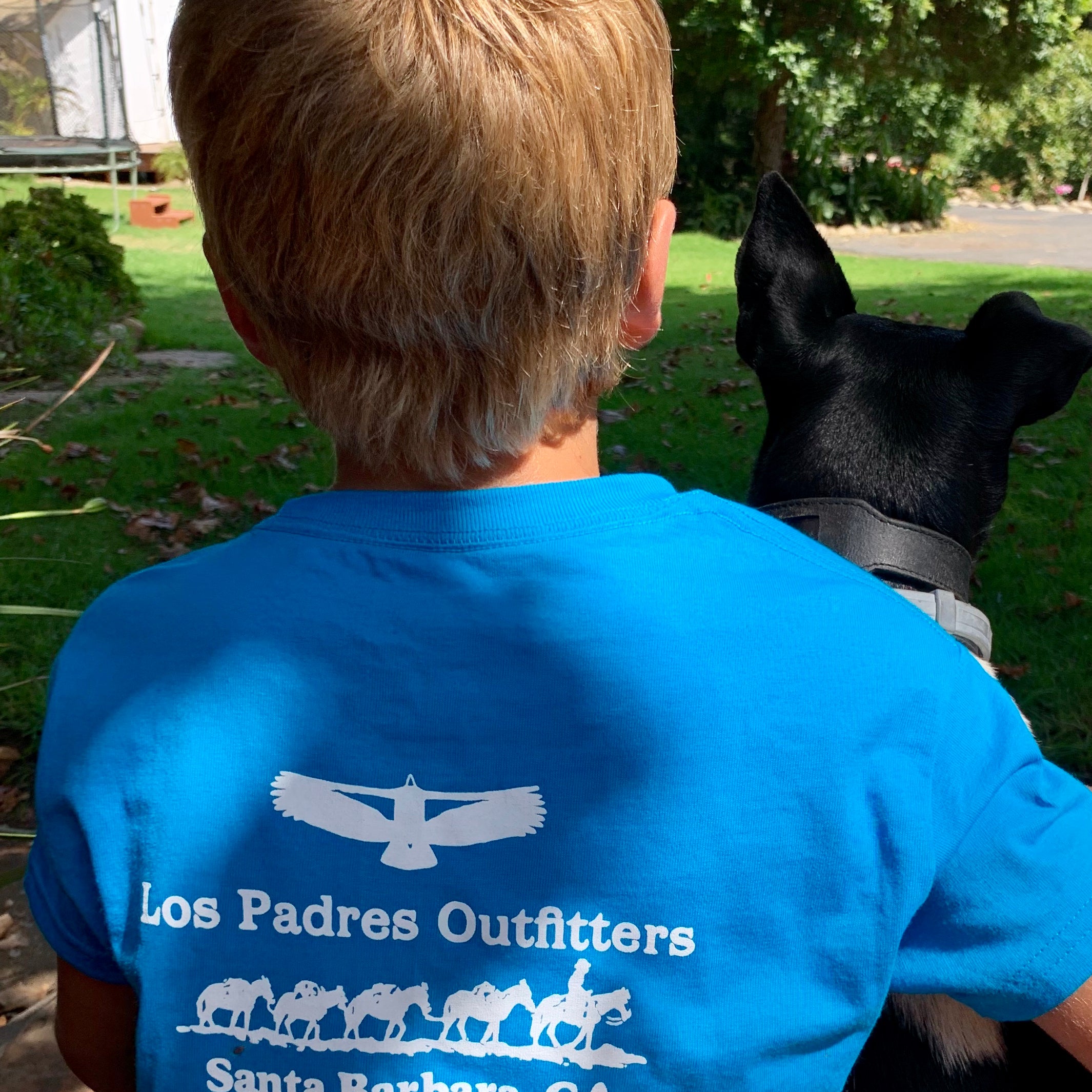 Back view of boy sitting outside wearing the comfortable blue cotton Kids Short Sleeve Tee with the Los Padres Outiftters logo on the back