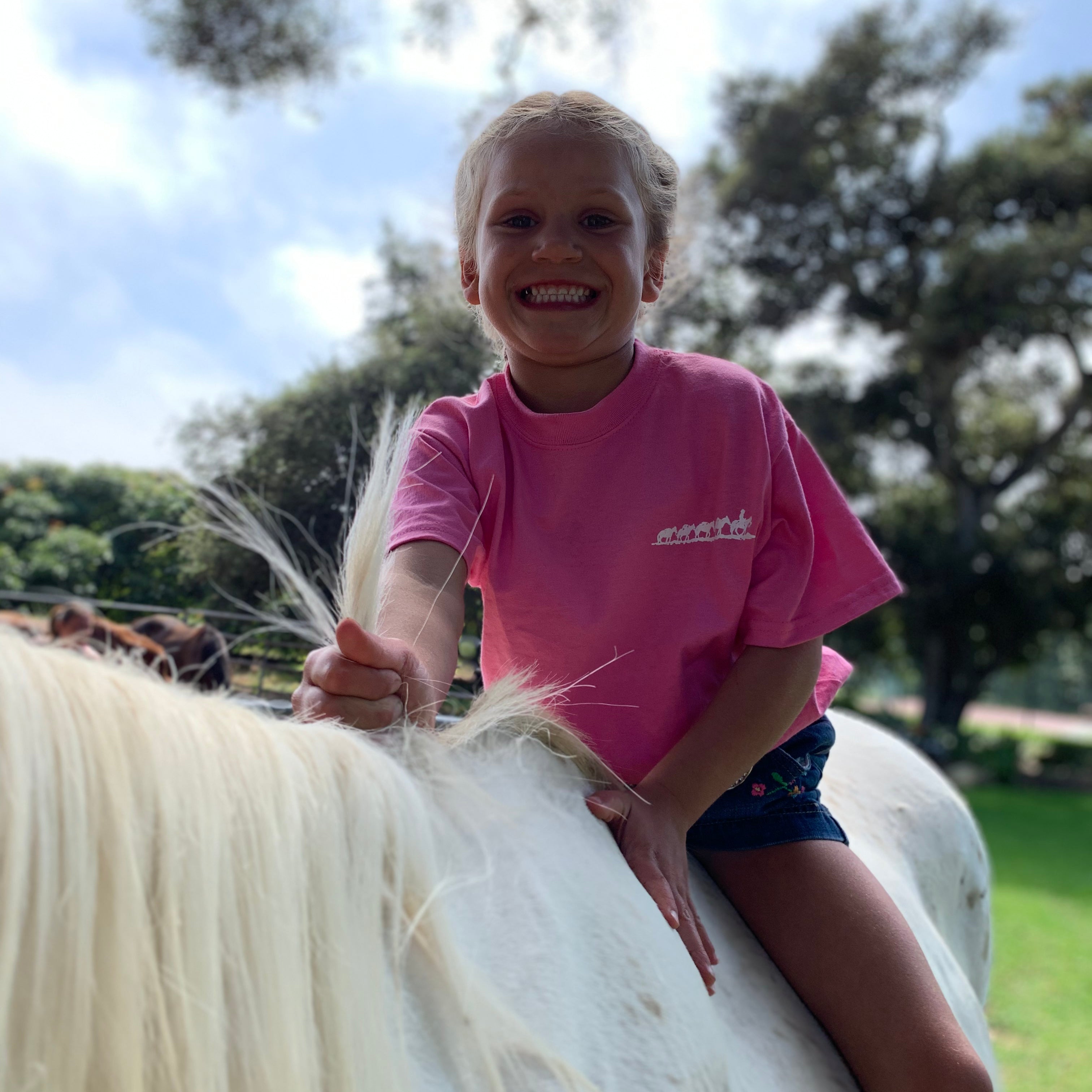 Front view of little girl riding a horse outside wearing the comfortable pink cotton Kids Short Sleeve Tee