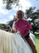 Front view of little girl riding a horse outside wearing the comfortable pink cotton Kids Short Sleeve Tee
